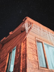 A wooden house corner with icicles hanging from the gutter against a backdrop of a starry night sky. The building is illuminated with a warm orange light, highlighting the texture of the wood and the clear window panes.