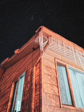 A wooden house corner with icicles hanging from the gutter against a backdrop of a starry night sky. The building is illuminated with a warm orange light, highlighting the texture of the wood and the clear window panes.