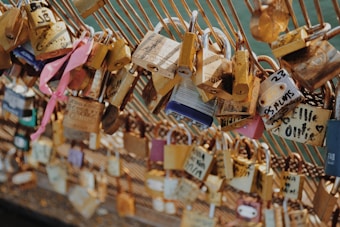 Multiple love locks are attached to a metal fence, each inscribed with personal messages or initials. The locks vary in size and color, with some decorated with ribbons, conveying personal or romantic commitments.