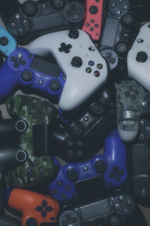 A collection of various gaming controllers placed haphazardly, featuring different styles and colors including white, black, red, blue, and camouflage patterns.