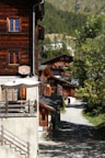 A mountain village scene featuring traditional wooden chalets nestled along a narrow pathway. The buildings are adorned with colorful flower boxes, and the area is enveloped by lush greenery and a backdrop of forested hills. The roofs and walls of the houses display rich wooden textures, and there are satellite dishes and outdoor seating on some terraces.