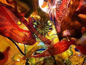 A vibrant, intricate display of colorful glass sculptures with wavy patterns and abstract forms. Light reflects through the glass, creating a kaleidoscope of vivid colors and shapes.