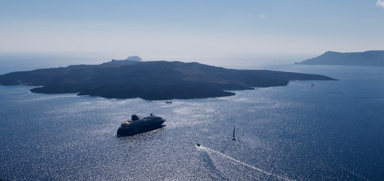 A large cruise ship is sailing in a vast, calm sea surrounded by scattered small islands. The water appears to shimmer under the sunlight, creating a serene and tranquil setting. A smaller sailboat is seen in the foreground, adding a sense of scale and contrast to the huge vessel. The distant horizon and soft haze suggest a peaceful, expansive seascape.