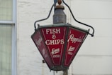 A vintage-style lamp with red panels showing the inscription 'Poppies Fish & Chips Since 1952'. The lamp hangs outside a building, and its design suggests a nostalgic or retro theme.