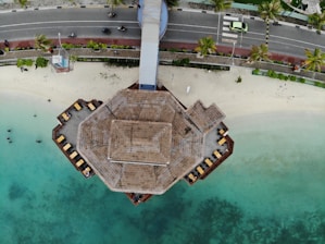 An aerial view of a hexagonal structure with a thatched roof extending over a turquoise body of water. The structure appears to be situated next to a sandy beach with palm trees lining a nearby road. Several people are visible on the deck, sitting on lounge chairs. A light green car and a few motorcycles are on the road.