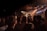 A live concert scene with a crowd of people gathered in front of a stage, watching a performance. Two performers are on stage, with one holding a microphone. Stage lights illuminate the area, creating a warm atmosphere. The background features the word 'MONAT' partially visible against a dotted light board.