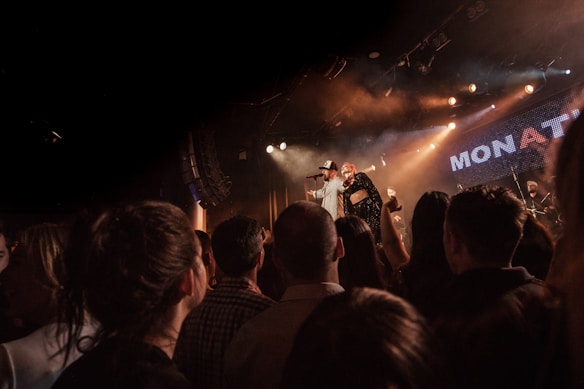 A live concert scene with a crowd of people gathered in front of a stage, watching a performance. Two performers are on stage, with one holding a microphone. Stage lights illuminate the area, creating a warm atmosphere. The background features the word 'MONAT' partially visible against a dotted light board.