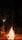A large bonfire is set up outdoors at night, surrounded by trees and a small structure in the background. The fire is built with logs stacked in a conical shape, with bright flames reaching upward and glowing embers floating in the dark sky. The ambient light from the fire casts a warm glow on the surroundings.