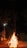 A large bonfire is set up outdoors at night, surrounded by trees and a small structure in the background. The fire is built with logs stacked in a conical shape, with bright flames reaching upward and glowing embers floating in the dark sky. The ambient light from the fire casts a warm glow on the surroundings.
