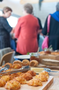 A table is filled with various baked goods, including scones or biscuits placed on a wooden tray. Behind, there are blurred silhouettes of people in conversation, dressed in casual clothing.