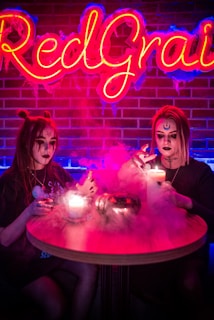 Two individuals are sitting at a round table with candles placed on it, surrounded by smoke. Both have face paint with gothic or mystical designs, creating an eerie atmosphere. A neon sign with the text 'Red Grail' is lit brightly in the background, adding to the vibrant setting.