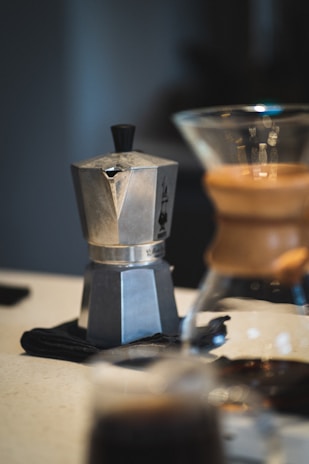 A stainless steel stovetop espresso maker is placed on a countertop, with a glass Chemex coffee maker in the foreground. The lighting is soft and creates a warm, inviting atmosphere.