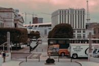 An urban scene featuring a street with a double-decker hop-on hop-off tour bus and a delivery truck. The background displays a mix of modern and classical architecture, including cranes and a tall, cylindrical building. There's a pedestrian walking on the sidewalk, and the area is surrounded by neatly trimmed trees.