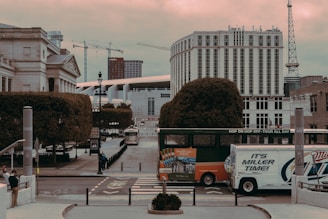 An urban scene featuring a street with a double-decker hop-on hop-off tour bus and a delivery truck. The background displays a mix of modern and classical architecture, including cranes and a tall, cylindrical building. There's a pedestrian walking on the sidewalk, and the area is surrounded by neatly trimmed trees.