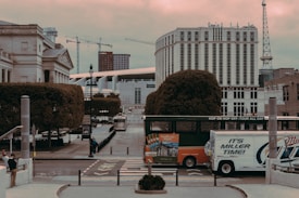 An urban scene featuring a street with a double-decker hop-on hop-off tour bus and a delivery truck. The background displays a mix of modern and classical architecture, including cranes and a tall, cylindrical building. There's a pedestrian walking on the sidewalk, and the area is surrounded by neatly trimmed trees.