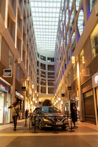 A modern indoor shopping mall with a high ceiling and glass roof, allowing natural light to illuminate the space. The architecture features clean lines and geometric designs. A car is displayed in the center, surrounded by a rope barrier. People are walking and browsing shops on either side, with store signs visible.