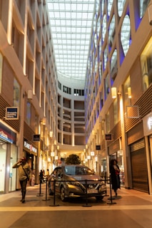 A modern indoor shopping mall with a high ceiling and glass roof, allowing natural light to illuminate the space. The architecture features clean lines and geometric designs. A car is displayed in the center, surrounded by a rope barrier. People are walking and browsing shops on either side, with store signs visible.