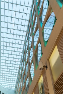 A modern architectural interior featuring a geometric design with large glass windows. The ceiling consists of a grid structure allowing natural light to illuminate the space. Walls display circles and diagonal lines, with a sleek and minimalistic design.