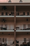 A three-story building facade featuring balconies with black metal railings. Each balcony is equipped with outdoor furniture, including chairs and tables, as well as potted plants and ceiling fans. The central vertical line of the building is made of exposed brick, contrasting with the white exterior walls.