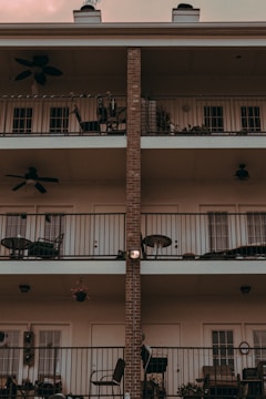 A three-story building facade featuring balconies with black metal railings. Each balcony is equipped with outdoor furniture, including chairs and tables, as well as potted plants and ceiling fans. The central vertical line of the building is made of exposed brick, contrasting with the white exterior walls.
