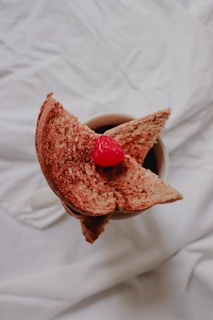 Two slices of toasted bread are arranged over a cup filled with a dark liquid, topped with a red cherry. The bread shows rich, toasted textures, and the cherry adds a pop of bright color.