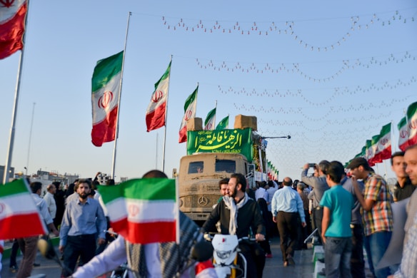 A crowd of people gathers on a street, many holding or surrounded by Iranian flags. The scene suggests a public gathering or parade, with a vehicle displaying a large banner written in a script language. Overhead, string lights are visible in the clear sky, enhancing the festive atmosphere.