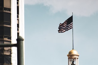 An American flag waves atop a tall flagpole on a building with a golden dome. A modern skyscraper with reflective windows is visible in the background against a clear sky.