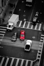 An aerial view of a busy intersection with several vehicles, including a prominent red car at the center. Pedestrians are visible on the sidewalk near a CVS pharmacy. The scene captures urban life with a mix of vehicles and people.
