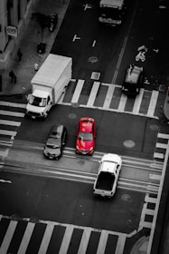 An aerial view of a busy intersection with several vehicles, including a prominent red car at the center. Pedestrians are visible on the sidewalk near a CVS pharmacy. The scene captures urban life with a mix of vehicles and people.