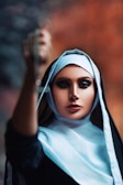 A woman wearing a black and white nun's habit stands with one arm extended forward, holding an object that is out of focus. Her makeup is dramatic, emphasizing her eyes, and the background features a blurred blend of dark and warm colors.