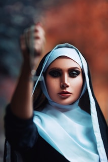 A woman wearing a black and white nun's habit stands with one arm extended forward, holding an object that is out of focus. Her makeup is dramatic, emphasizing her eyes, and the background features a blurred blend of dark and warm colors.