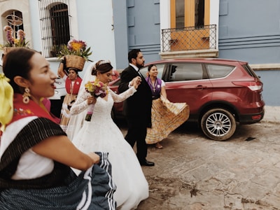 A wedding procession takes place on a cobblestone street with a bride in a white gown holding a bouquet, and a groom in a dark suit. They are surrounded by individuals dressed in traditional attire, with a woman carrying a basket of colorful flowers on her head.