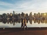 two people walking near body of water viewing city with high-rise buildings under white and blue sky during daytime