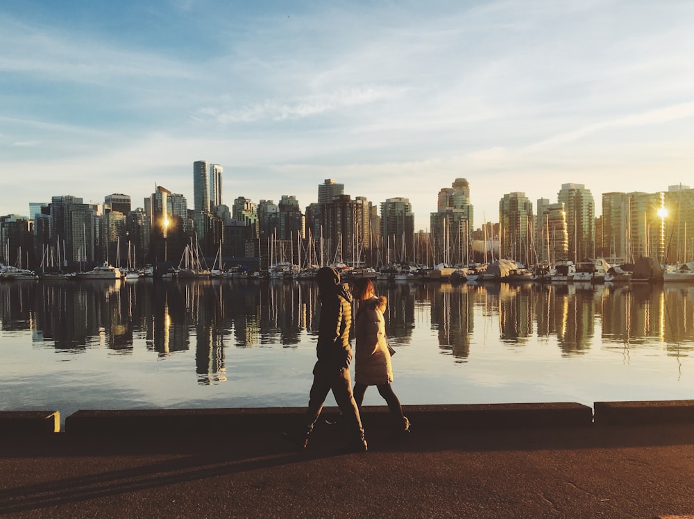 two people walking near body of water viewing city with high-rise buildings under white and blue sky during daytime