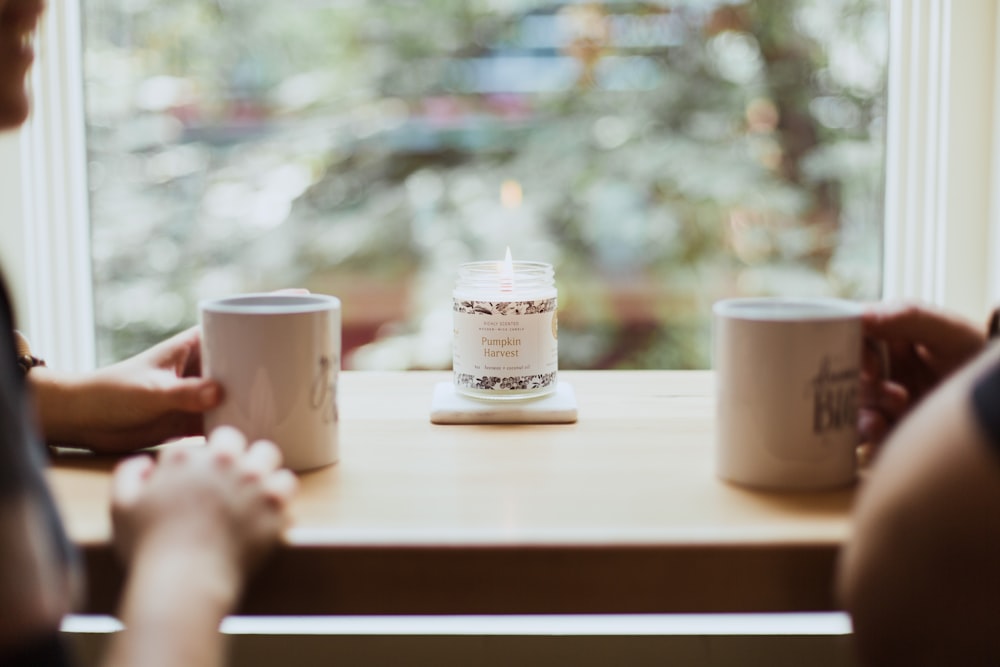 two person holding white mugs on table front of window