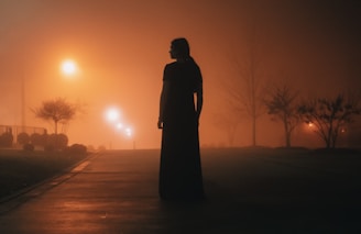 A shadowy figure standing at the edge of a fog-covered forest path at twilight.