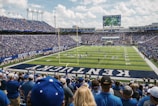 A large football stadium filled with fans watching a live game. Players are on the field, and the atmosphere is lively with a full audience. The scoreboard displays the action, and banners for the Kentucky Wildcats are visible.