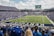 A large football stadium filled with fans watching a live game. Players are on the field, and the atmosphere is lively with a full audience. The scoreboard displays the action, and banners for the Kentucky Wildcats are visible.