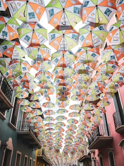 Multiple colorful umbrellas are suspended in rows above a narrow street, creating a vibrant canopy. The umbrellas display various illustrations and text, adding a lively and artistic atmosphere to the urban setting. The buildings on either side feature balconies and are painted in soft pastel colors, enhancing the charming aesthetic.