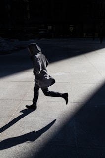 A person is running across a concrete surface with long shadows stretching behind. The surroundings are dimly lit, with strong contrasts of light and darkness creating a dramatic effect.