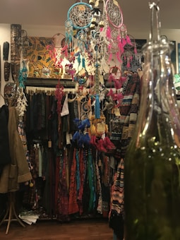A colorful display in a shop features hanging dreamcatchers with feathers in vibrant pinks, blues, and reds. Below the dreamcatchers, a variety of patterned clothes hang neatly on racks. The setting is adorned with eclectic decorations, including masks and artwork on the walls, creating a bohemian atmosphere. A large green bottle is prominently visible in the foreground.