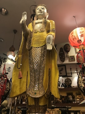 A tall, ornate statue of a standing figure with a serene expression and eyes closed. The statue is adorned in intricate gold garments with a mesh-like pattern and holds a long bead necklace in one hand. The surrounding area is filled with various cultural artifacts, including a red paper lantern with Chinese characters, other decorative items, and art pieces on the wall.