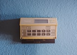 A vintage Honeywell thermostat is mounted on a textured light blue wall. The thermostat features a beige color with numerous buttons for programming and setting temperatures, along with a small display screen.
