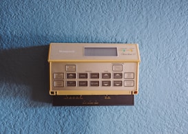 A vintage Honeywell thermostat is mounted on a textured light blue wall. The thermostat features a beige color with numerous buttons for programming and setting temperatures, along with a small display screen.