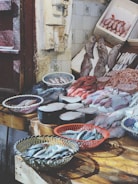 A fish market display features a variety of fresh seafood arranged on wooden tables. Baskets hold small fish, while large cuts of fish meat and whole fish are laid out neatly. The rustic setting includes tiled walls and weathered doors.