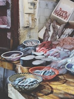 A fish market display features a variety of fresh seafood arranged on wooden tables. Baskets hold small fish, while large cuts of fish meat and whole fish are laid out neatly. The rustic setting includes tiled walls and weathered doors.