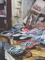 A fish market display features a variety of fresh seafood arranged on wooden tables. Baskets hold small fish, while large cuts of fish meat and whole fish are laid out neatly. The rustic setting includes tiled walls and weathered doors.