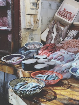A fish market display features a variety of fresh seafood arranged on wooden tables. Baskets hold small fish, while large cuts of fish meat and whole fish are laid out neatly. The rustic setting includes tiled walls and weathered doors.