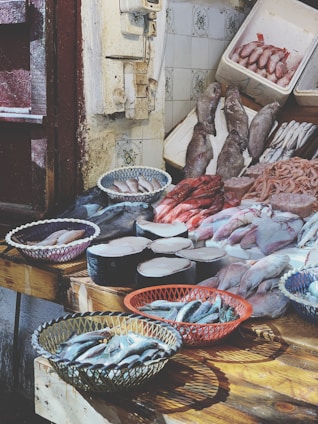 A fish market display features a variety of fresh seafood arranged on wooden tables. Baskets hold small fish, while large cuts of fish meat and whole fish are laid out neatly. The rustic setting includes tiled walls and weathered doors.