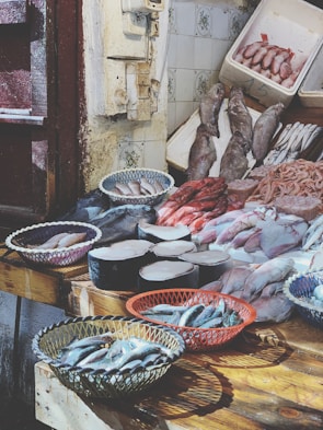 A fish market display features a variety of fresh seafood arranged on wooden tables. Baskets hold small fish, while large cuts of fish meat and whole fish are laid out neatly. The rustic setting includes tiled walls and weathered doors.